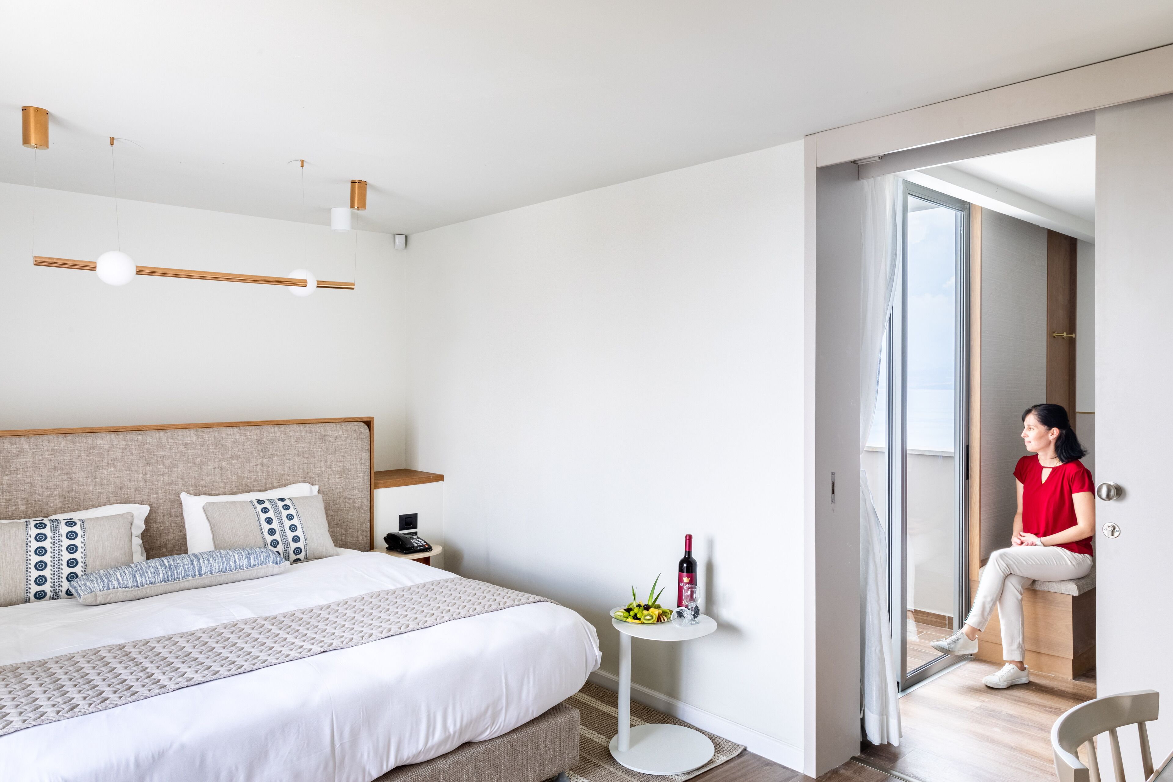 A modern hotel room with a large bed, a small round table with a bottle and fruit, and a view through an open door.