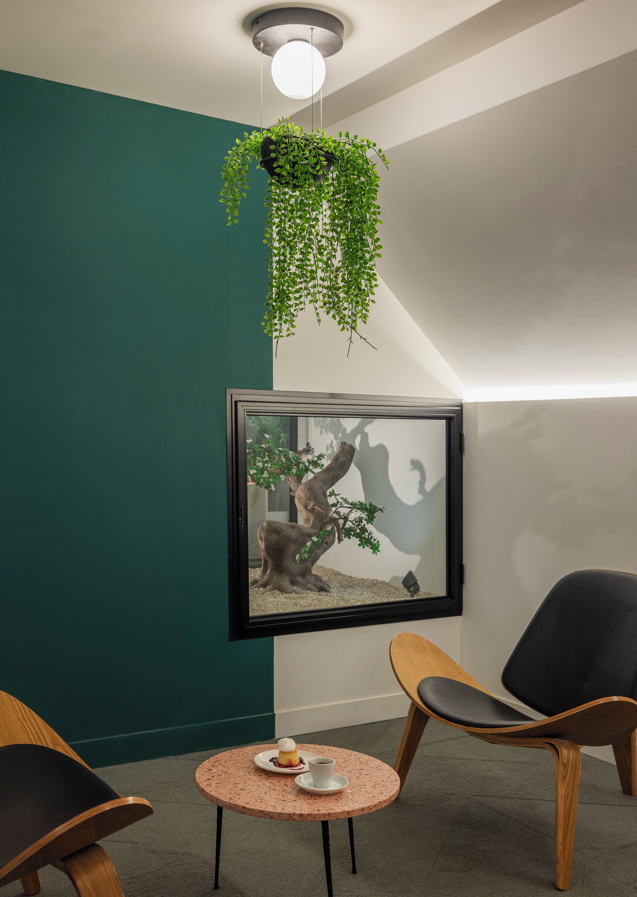Een kamer met een ingelijste bonsai, twee stoelen, een klein tafeltje met een kopje en gebak, en een hangplant.