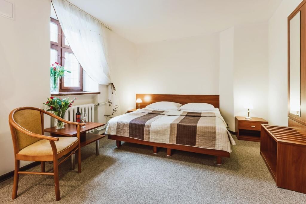Quarto com cama de madeira, cadeira de madeira, mesa pequena, radiador, janela com cortinas e mesa de cabeceira com abajur.