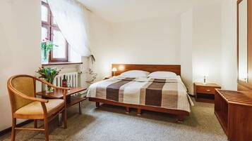 Quarto com cama de madeira, cadeira de madeira, mesa pequena, radiador, janela com cortinas e mesa de cabeceira com abajur.