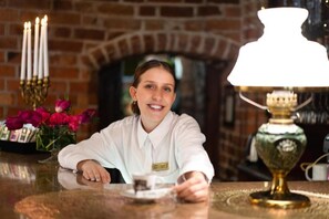 Una persona sentada a una mesa con un gafete, un candelabro con velas encendidas y una lámpara con pantalla blanca.