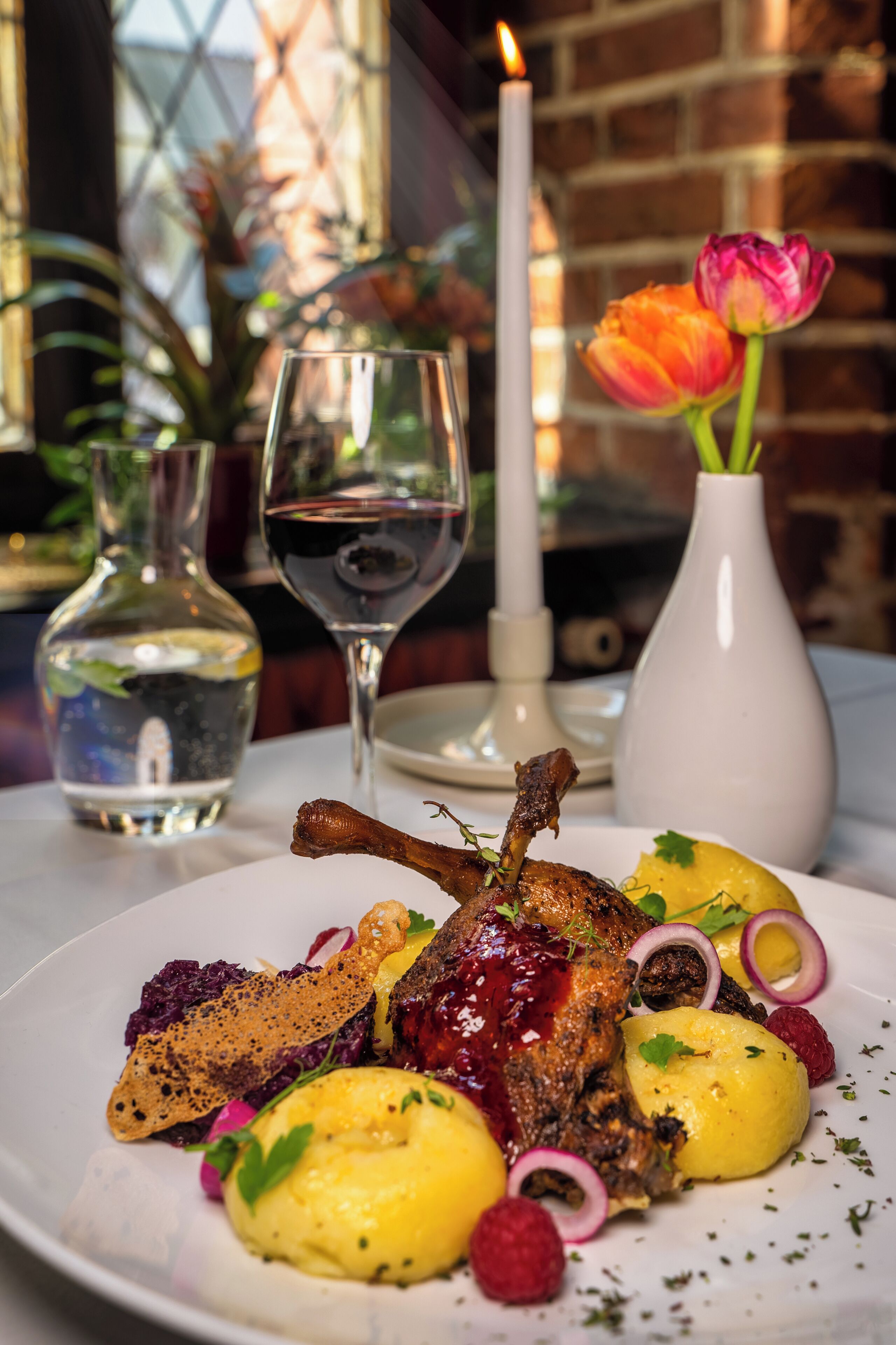 A plate of food with a lamb shank, potatoes, and berries, accompanied by a glass of wine and a candle.