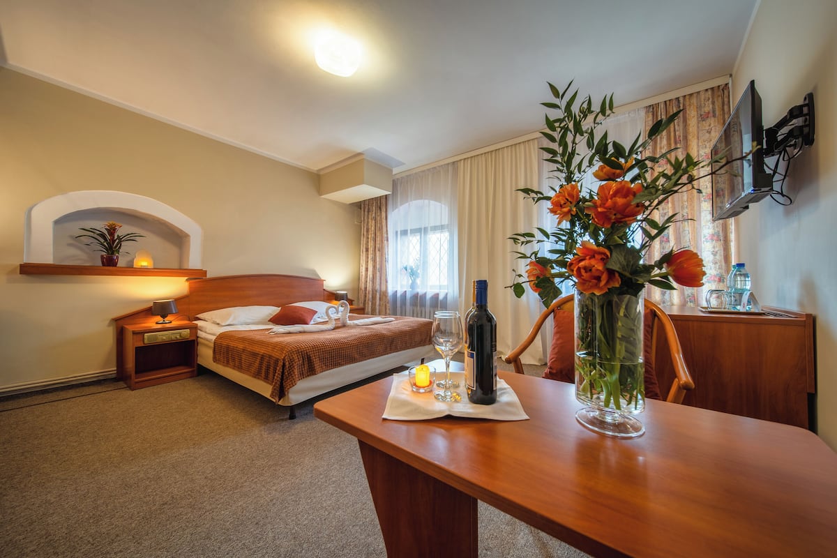 A hotel room with a bed, bedside table with a lamp, a small table with a vase of flowers, a bottle of wine, and a TV mounted on the wall.