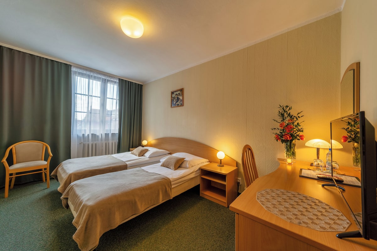 A hotel room with two beds, a desk with a computer, a chair, a small table with a vase of flowers, and a window with curtains.