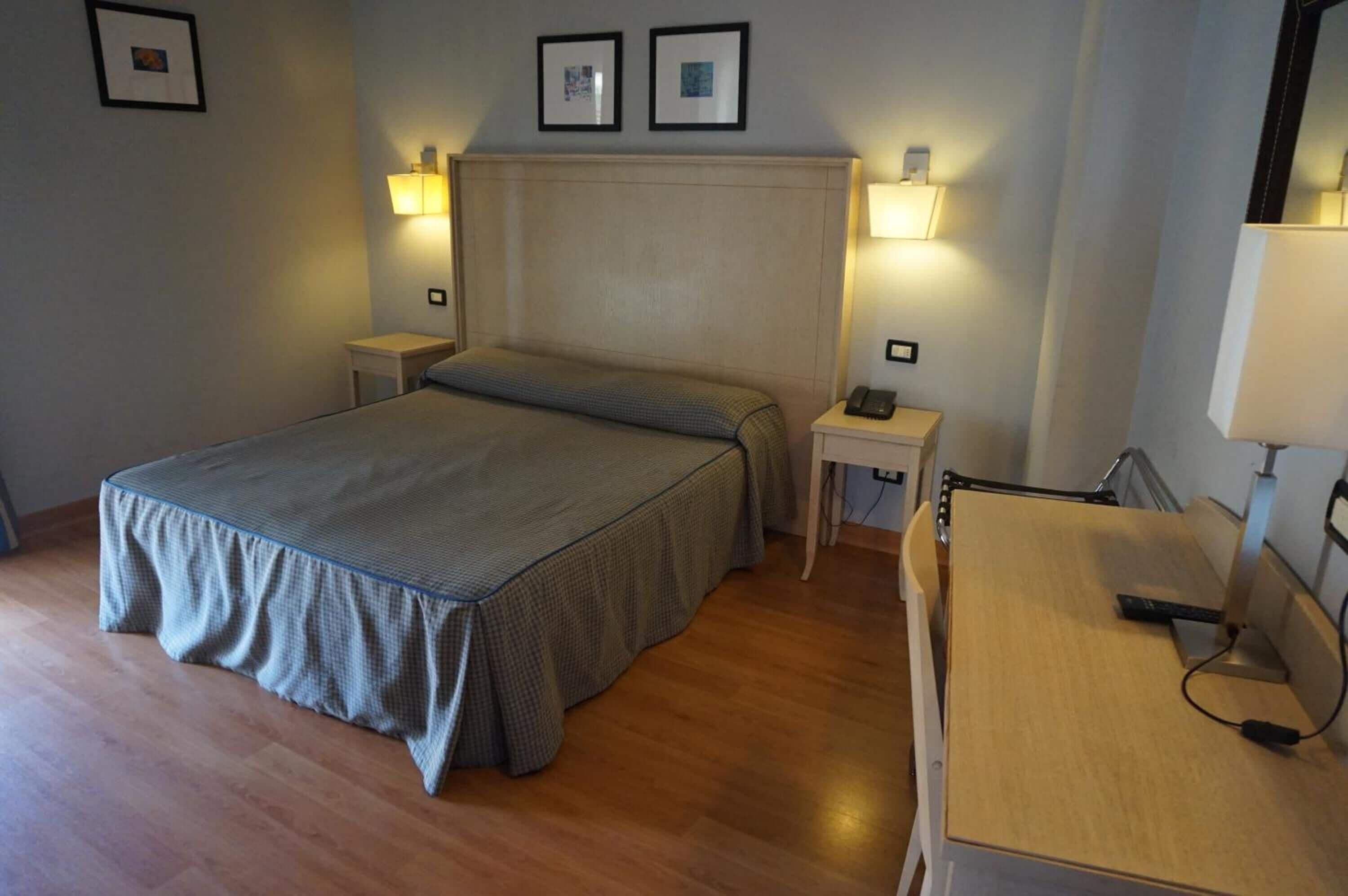 Una habitación de hotel con cama, mesitas de noche, un escritorio y lámparas de pared.