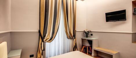A hotel room with a bed, a desk with a chair, a TV mounted on the wall, and a window with curtains.