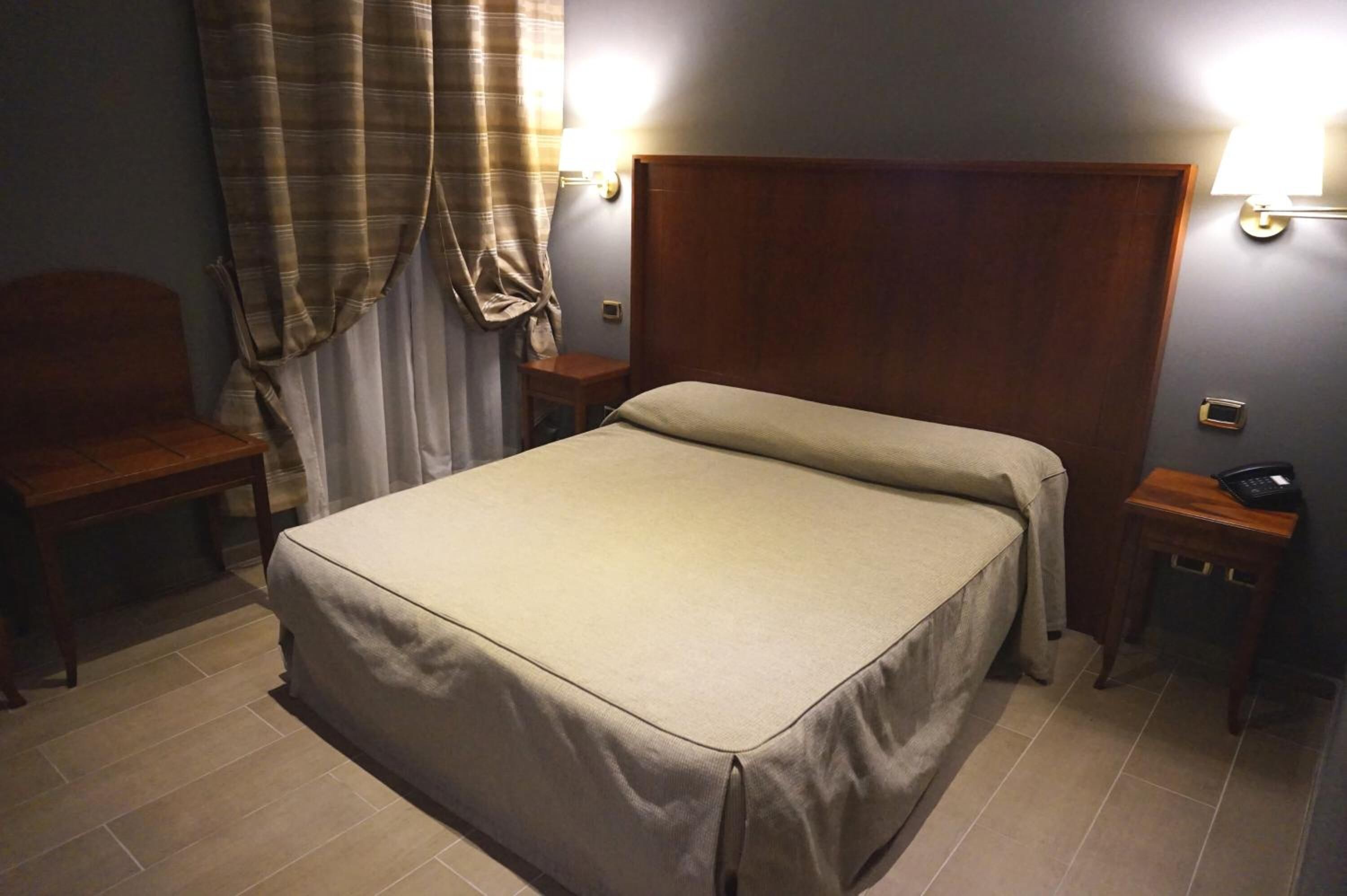 Une chambre d’hôtel avec un lit, deux tables de chevet, une chaise, une lampe et un rideau.