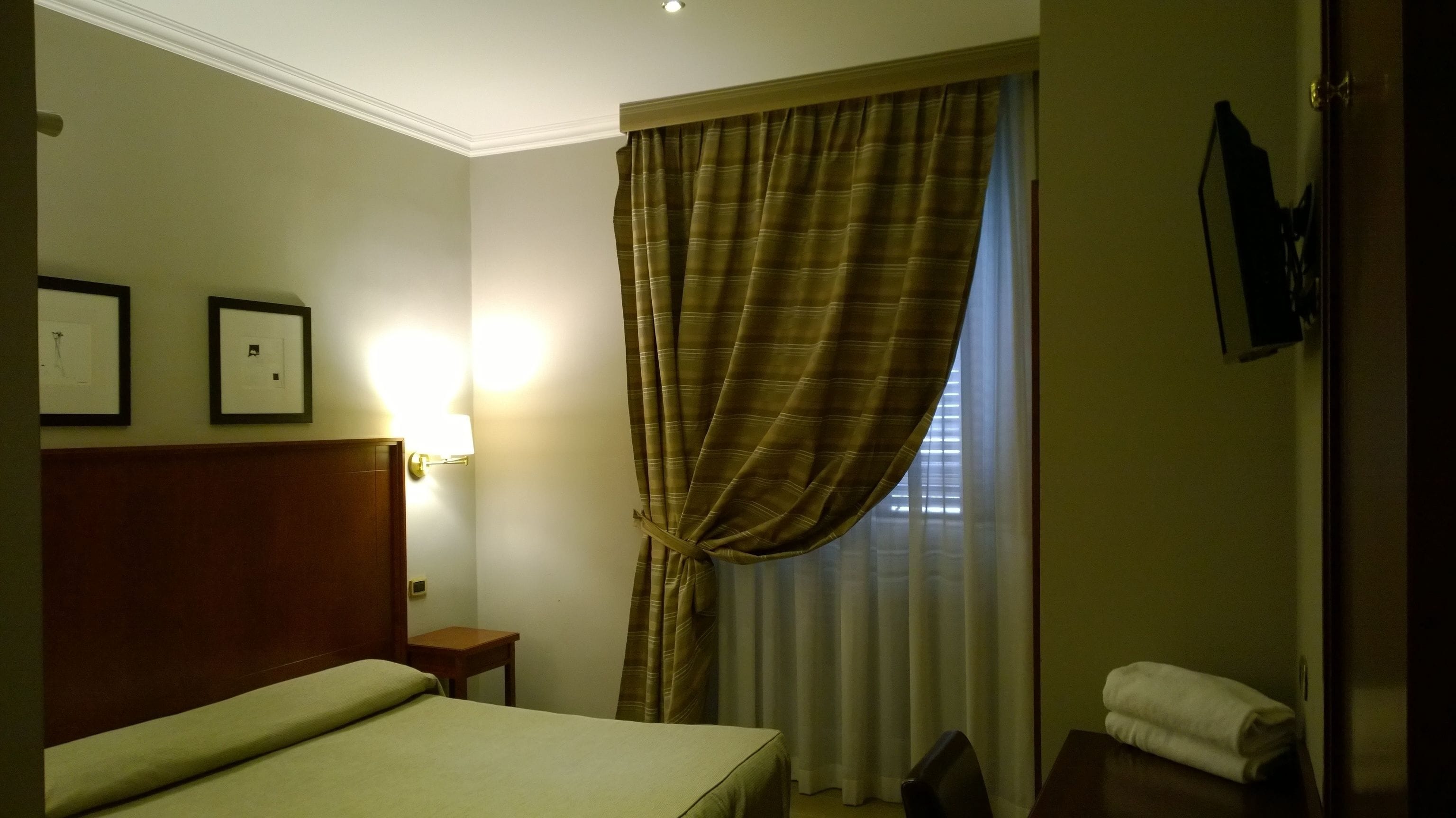 Habitación de hotel con cama, mesita de noche, televisor montado en la pared y ventana con cortinas.