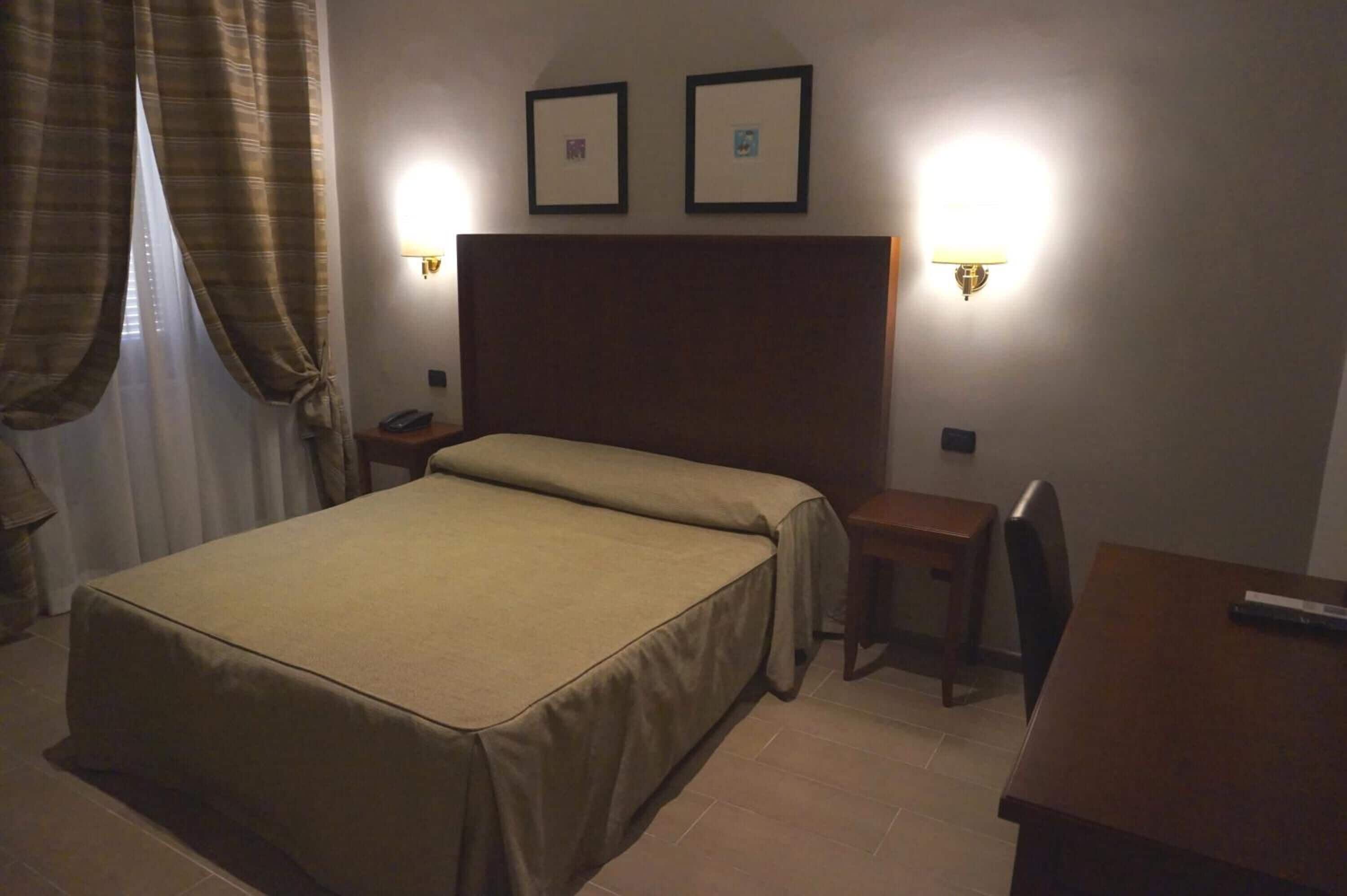 A hotel room with a bed, bedside table, chair, and two wall-mounted lamps.