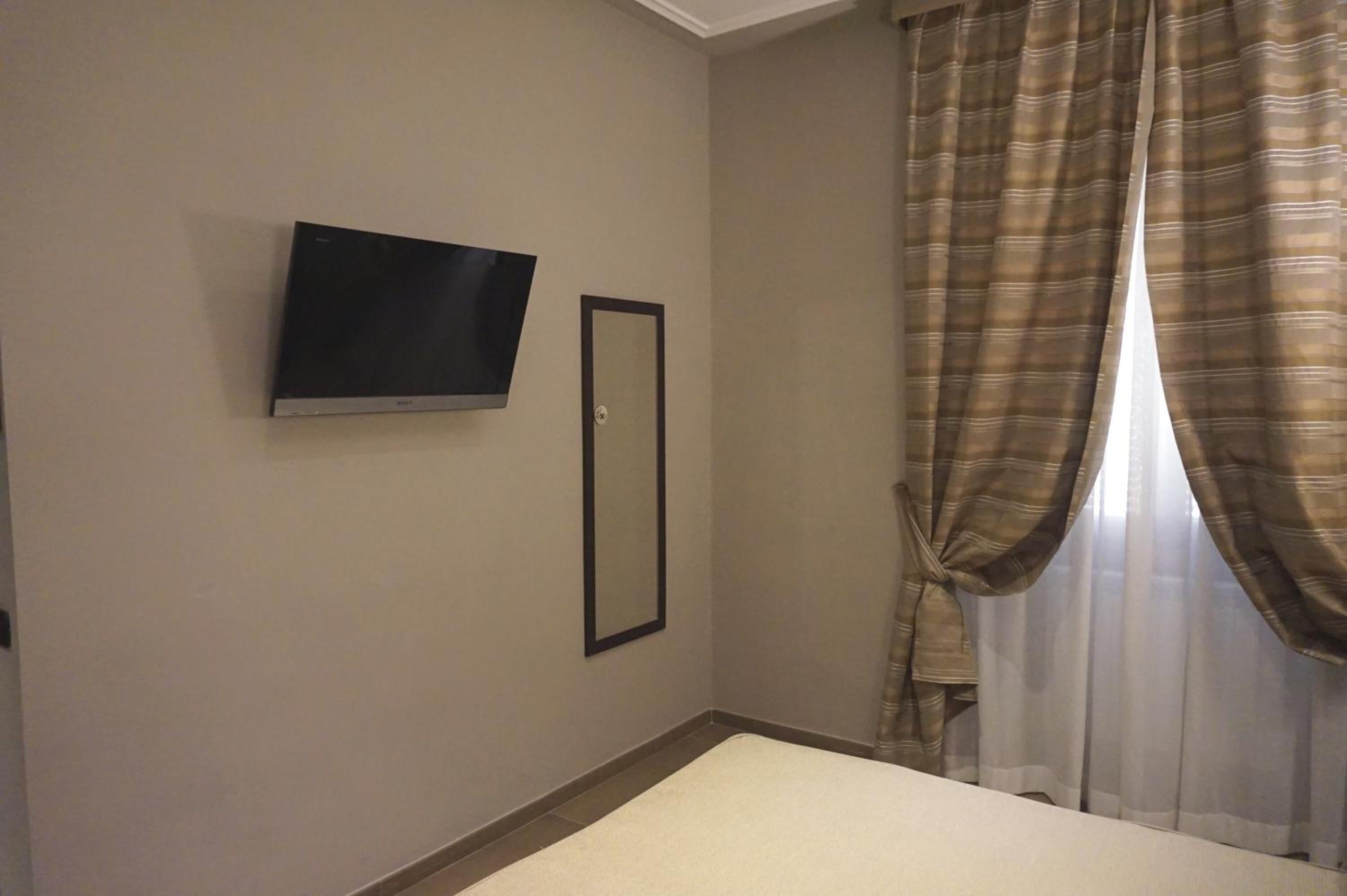 Quarto de hotel com televisão de tela plana na parede, espelho emoldurado e janela com cortinas xadrez.