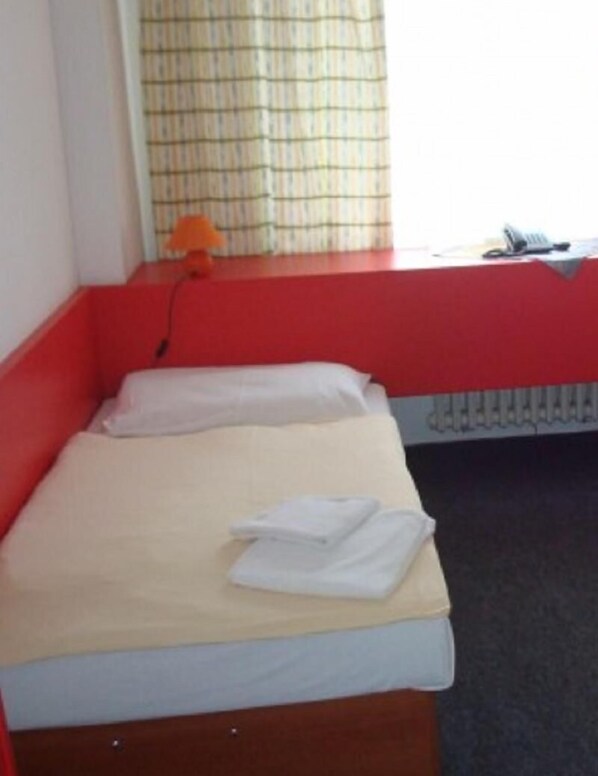 A small, single-bed room with a red lower half and a beige upper half. There is a window with a checkered curtain, a radiator, and a small orange toy on the window sill. - Slavia (Prague)