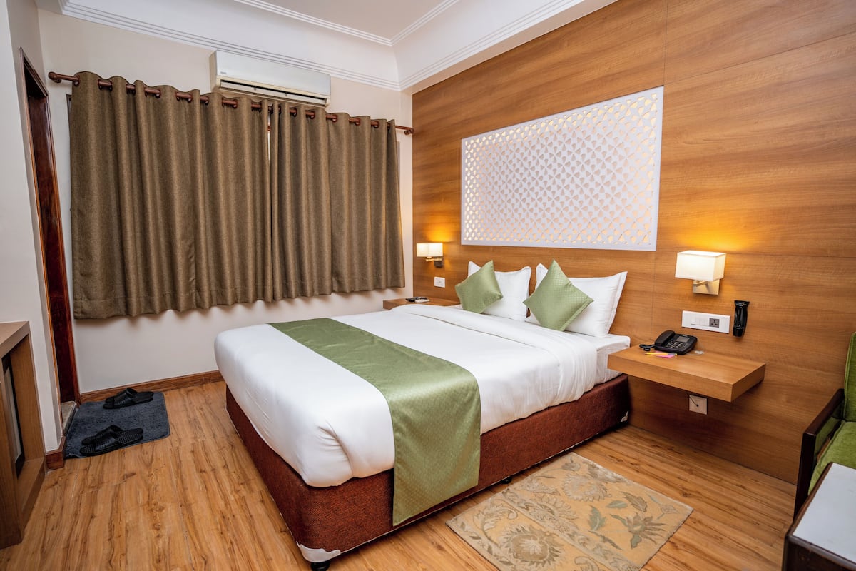 A hotel room with a wooden headboard, a bed with white and green bedding, a bedside table with a lamp, and a patterned wall decoration.