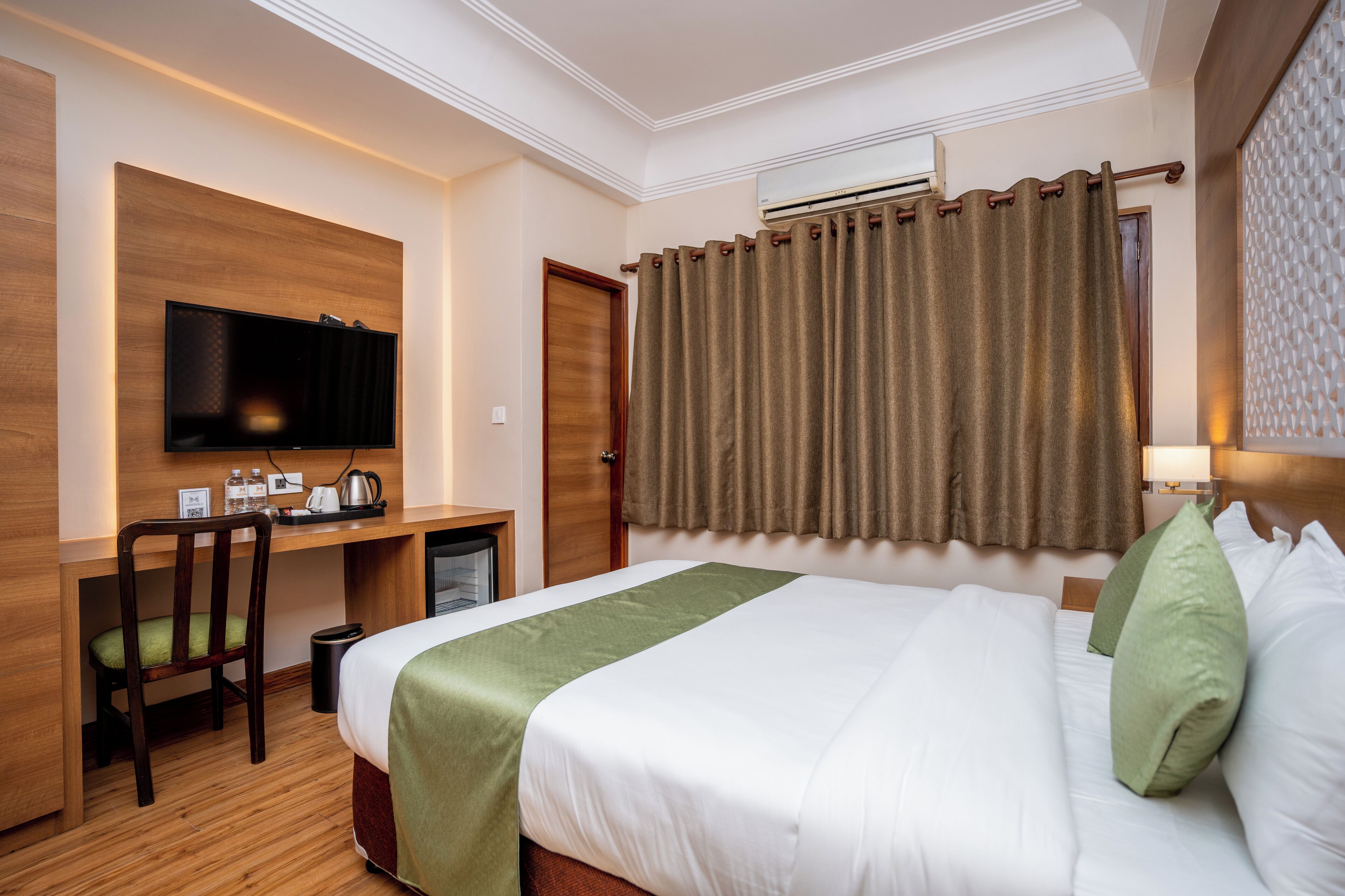 A hotel room with a bed, a desk with a chair, a television, and an air conditioning unit.