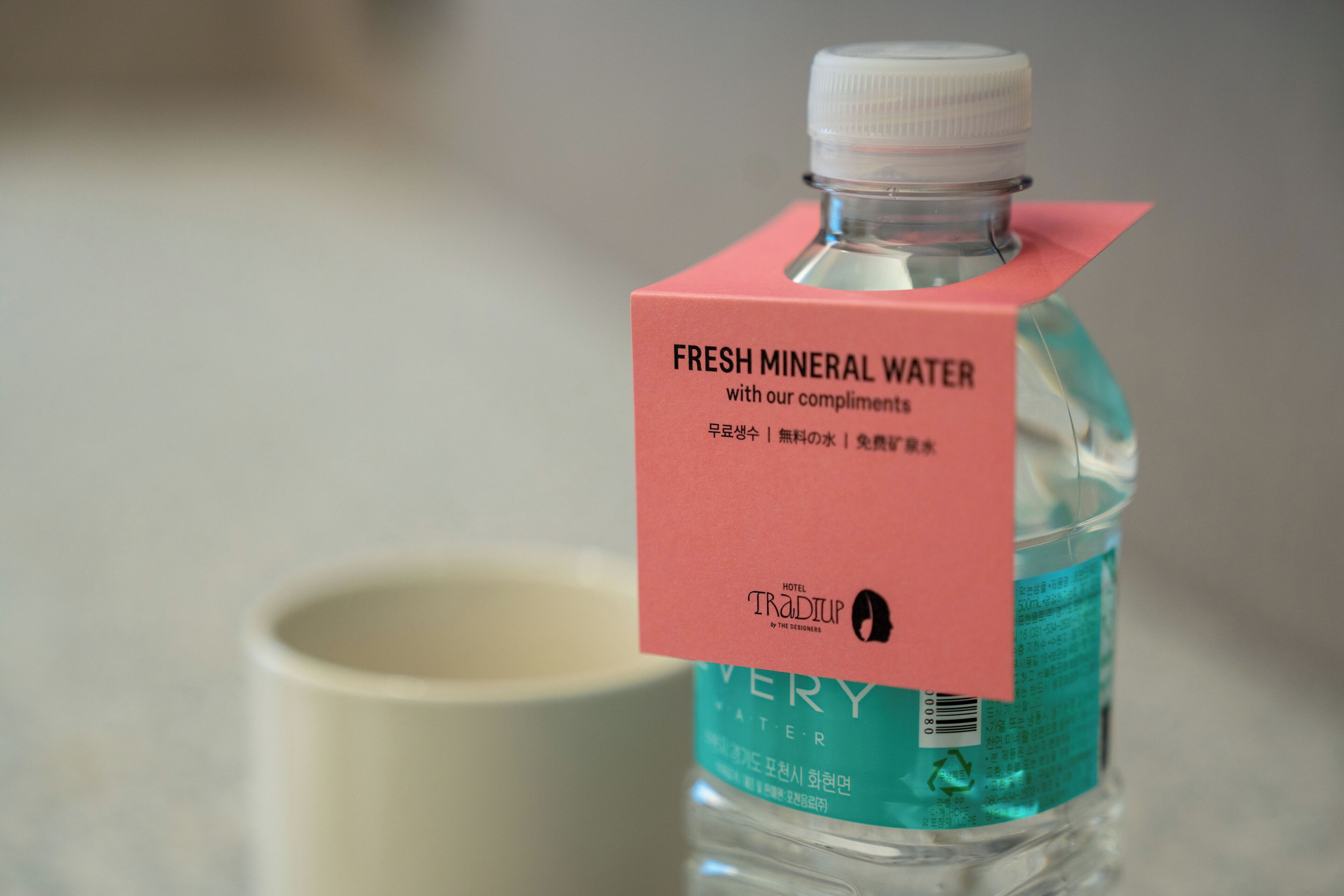 Eine Wasserflasche mit rosa Etikett und weißem Verschluss, neben einem weißen Becher platziert.