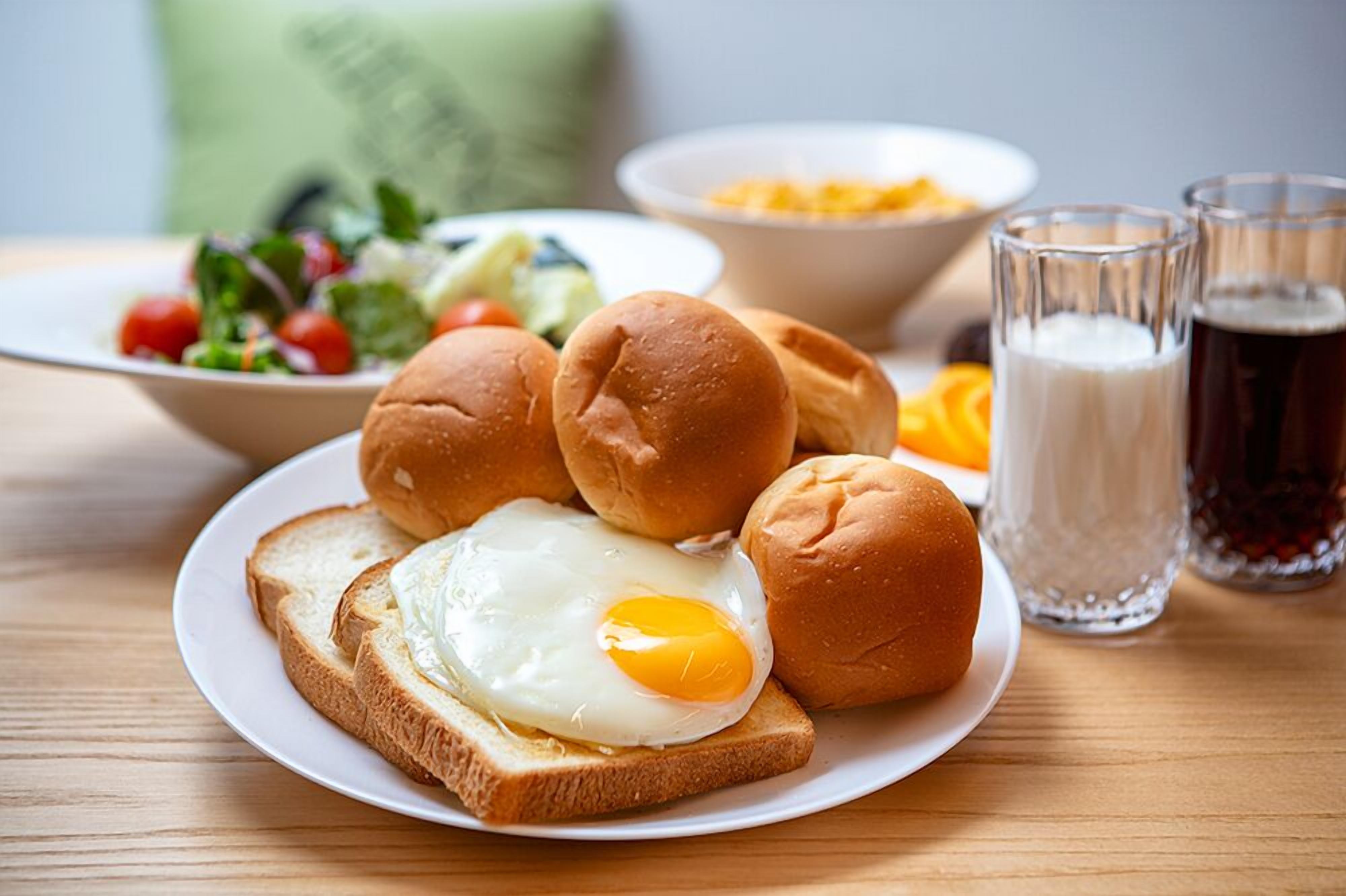 Un plato con un huevo frito sobre tostada, un bol de ensalada y dos vasos de leche junto con una bebida oscura, todo sobre una mesa de madera.