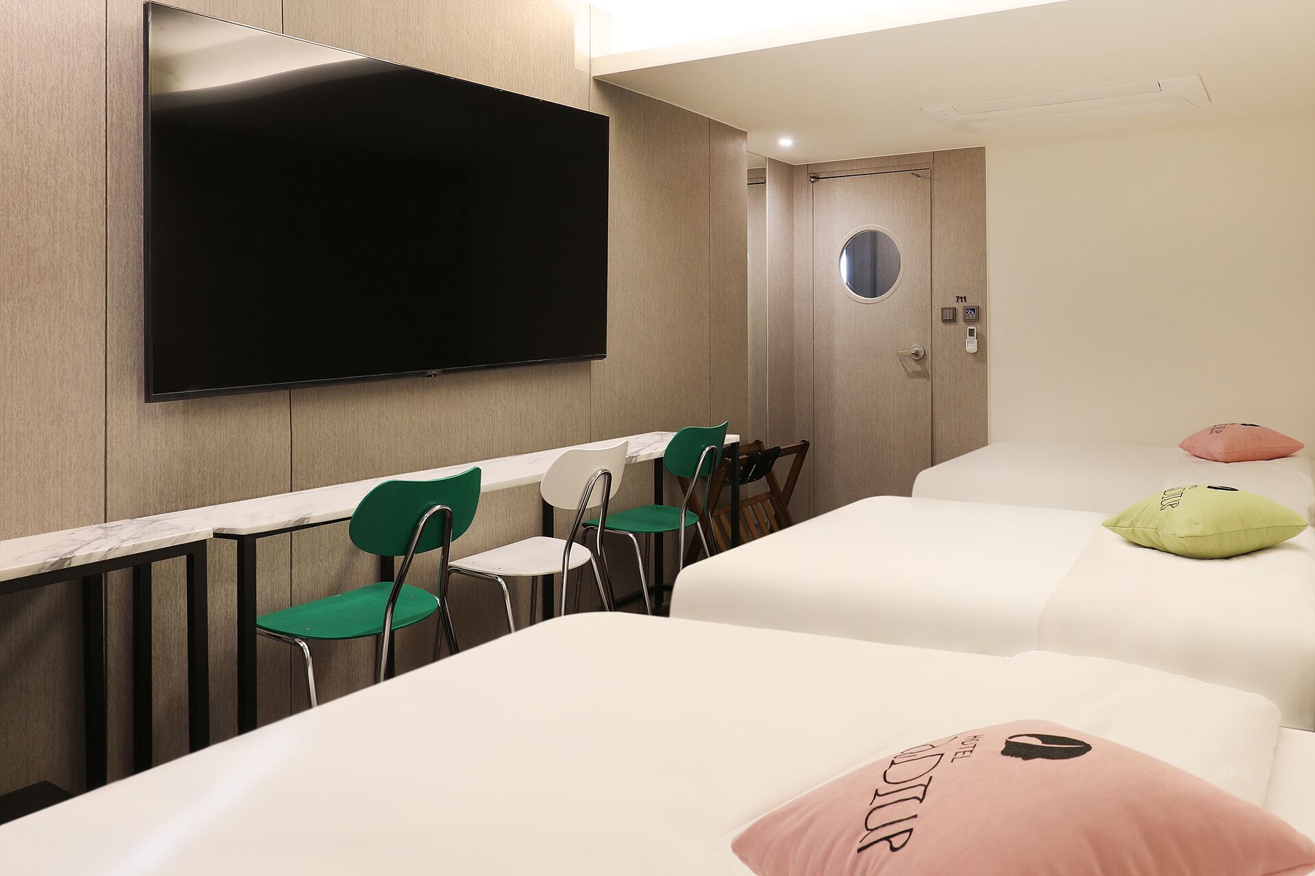 Habitación de hotel con televisión de pantalla plana montada en la pared, cama con almohada rosa, silla verde y una mesita.