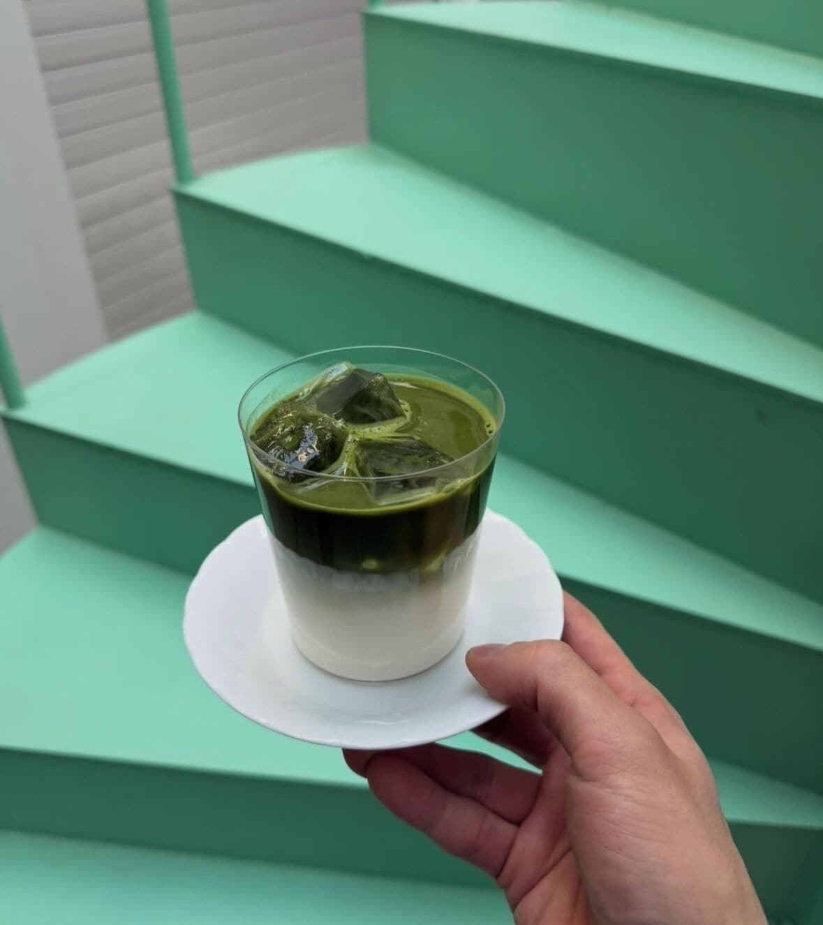Uma mão segurando uma chávena de matcha sobre um pires branco, com degraus verdes ao fundo.