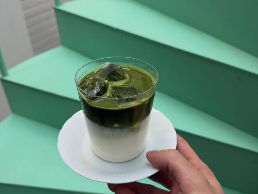 Uma mão segurando uma chávena de matcha sobre um pires branco, com degraus verdes ao fundo.