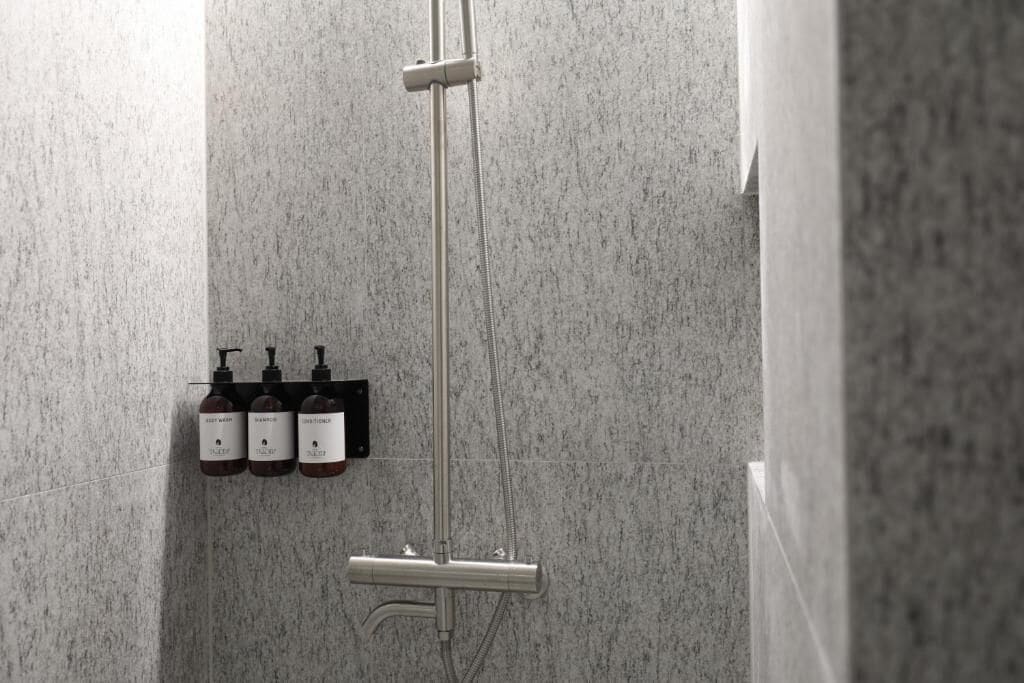 Une douche équipée d’un pommeau de douche à main et d’un robinet mural. On trouve trois bouteilles de shampoing et d’après-shampoing sur une étagère.