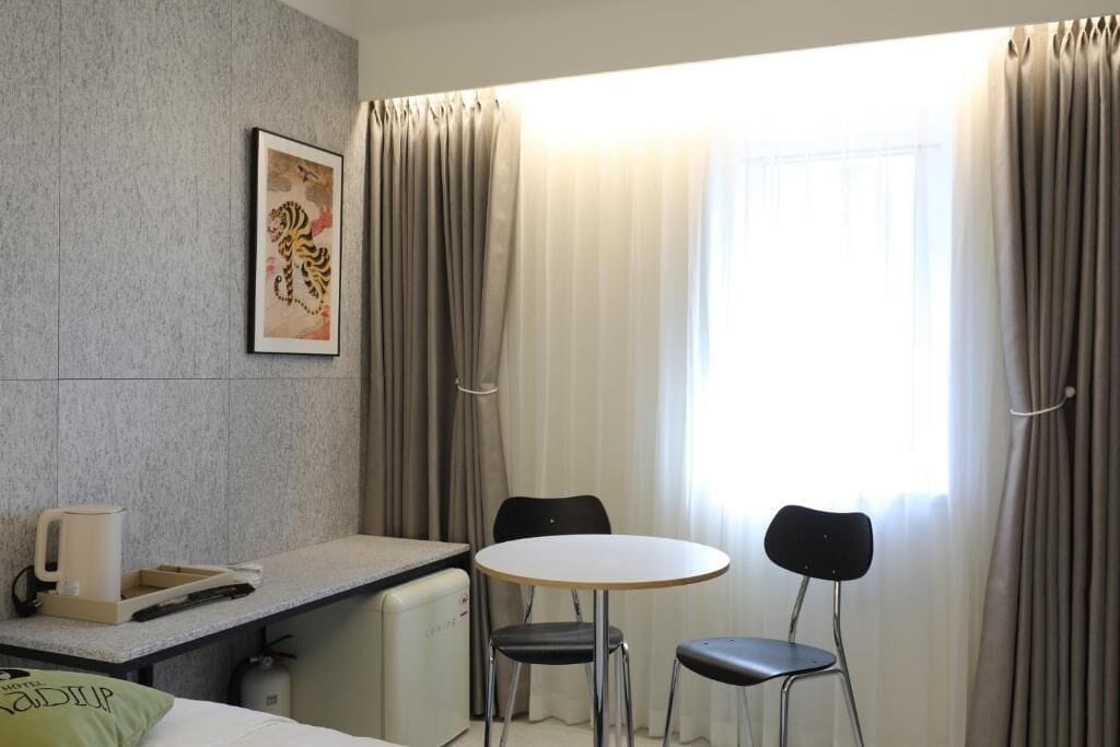 Habitación de hotel con cama, escritorio y silla, una mesita con dos sillas, un cuadro en la pared y una ventana con cortinas.