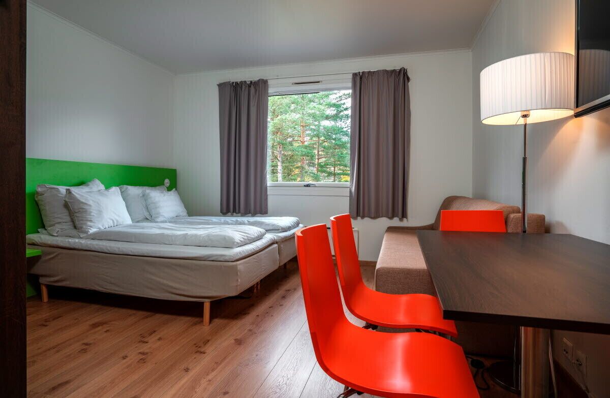 Una habitación con una cama, dos sillas rojas, una mesa y una lámpara.