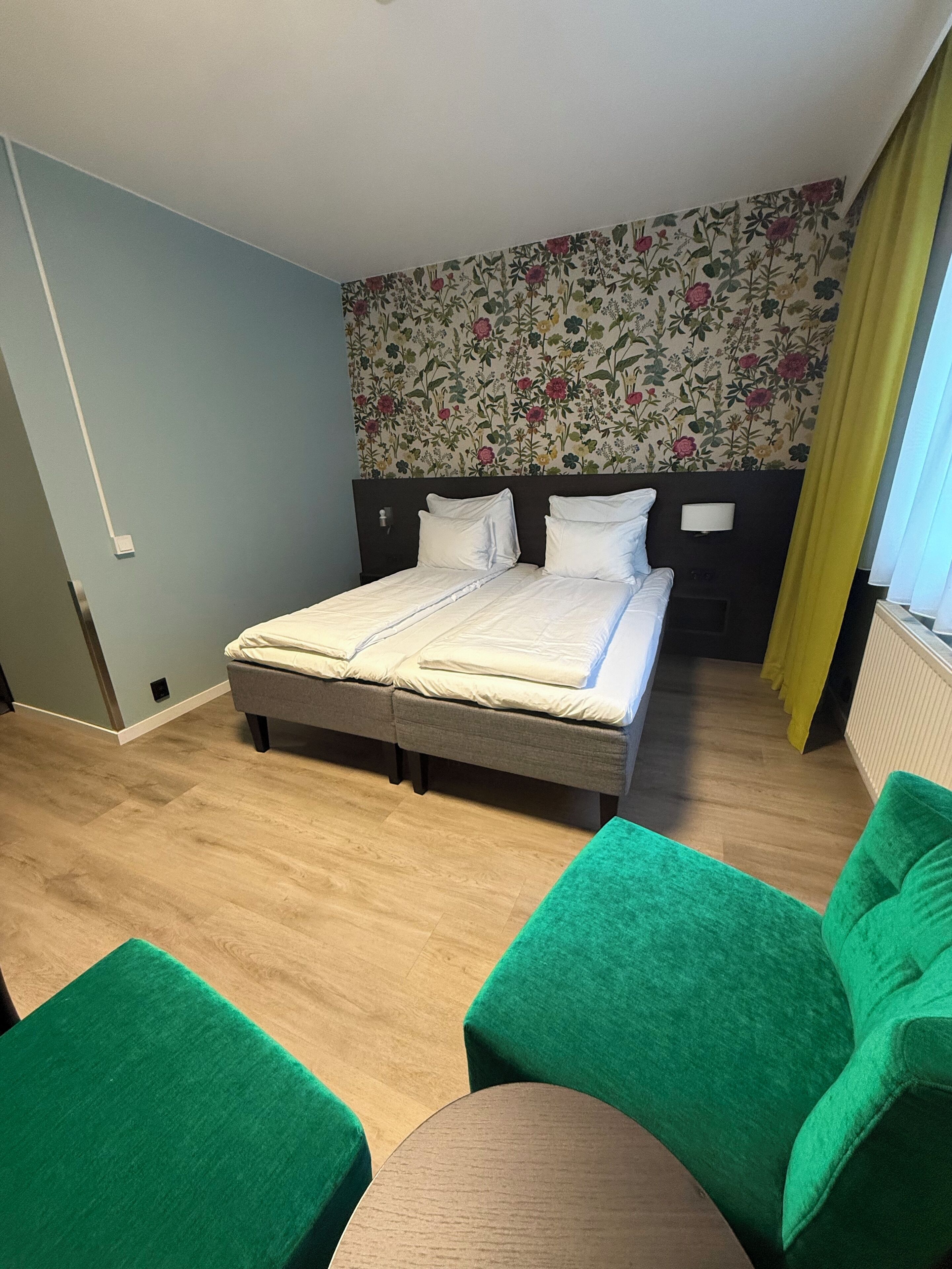 A hotel room with a bed, green armchairs, a small table, floral wallpaper, and a window with curtains.