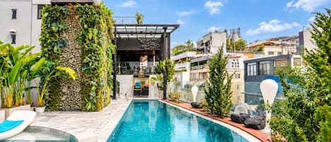 A swimming pool with a stone wall and greenery on one side, and a bar area with a view of the cityscape in the background.
