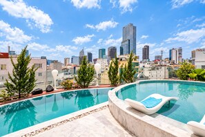 Piscina infinita en la azotea con vista al horizonte de la ciudad, rodeada de plantas en macetas y una silla de descanso.