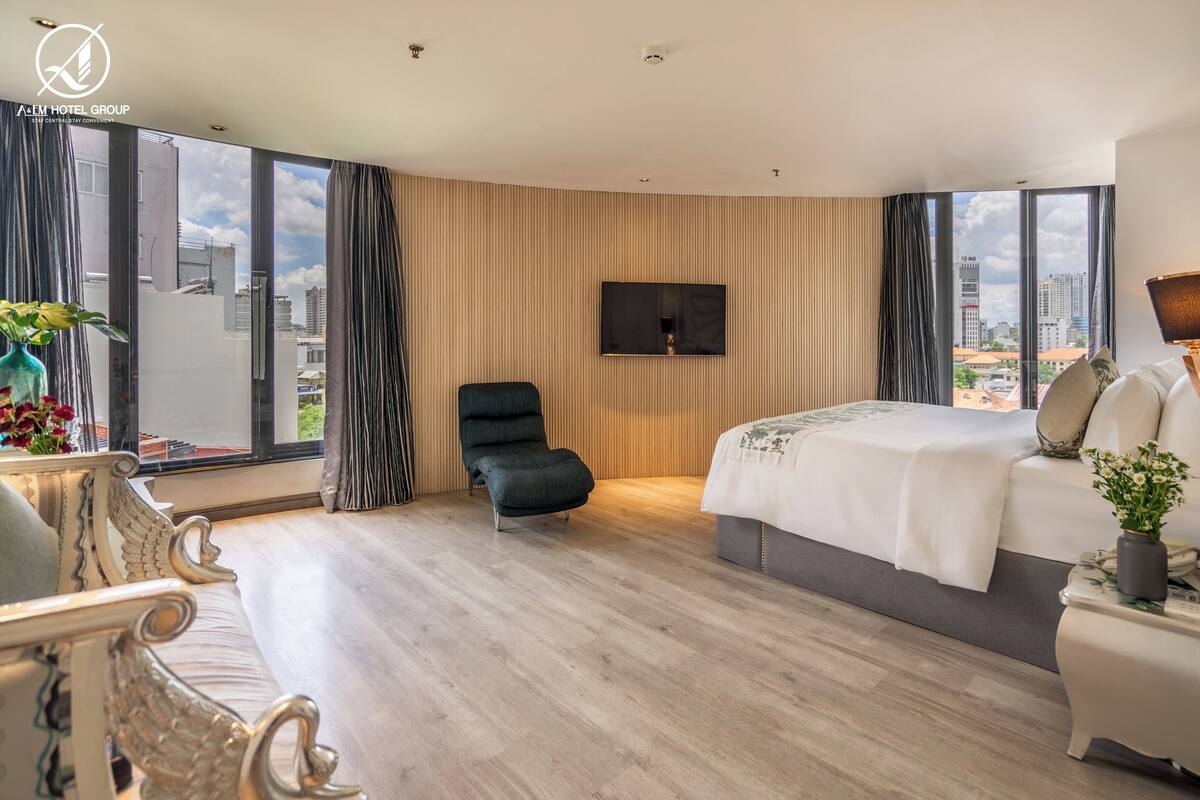 Habitación de hotel moderna con una cama grande, zona de estar con una silla, televisor montado en la pared y vistas a la ciudad a través de amplios ventanales.