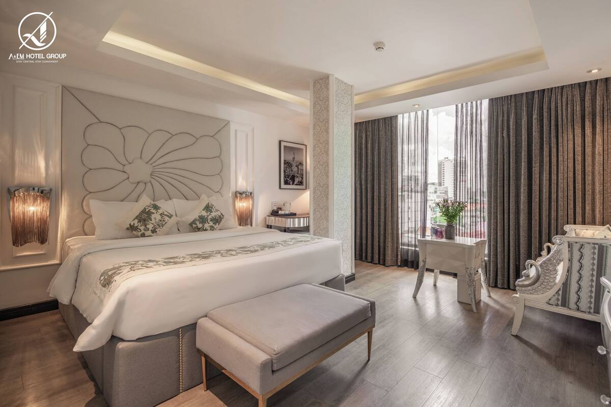 Amplia habitación de hotel con una cama grande, un área de descanso con una mesita y sillas, y vista a la ciudad a través de amplios ventanales.