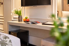 Une chambre d’hôtel moderne avec un téléviseur à écran plat fixé au-dessus d’un bureau en bois, un vase avec des fleurs blanches, un saladier de fruits et un magazine.