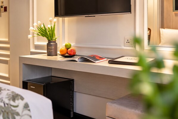 Ein modernes Hotelzimmer mit einem flachbildfernseher an der Wand über einem Holztisch, einer Vase mit weißen Blumen, einer Obstschale und einer Zeitschrift.
