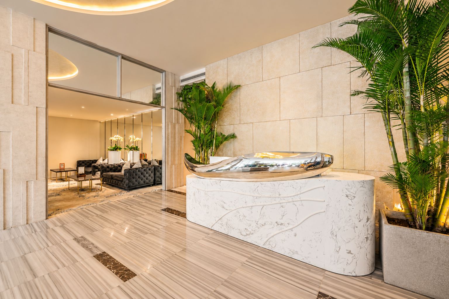 A modern lobby with a marble reception desk, a seating area with a sofa and chairs, and potted plants.