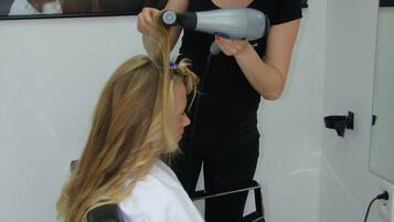 A salon interior with a client seated in a chair, a stylist using a hairdryer, and a poster on the wall.