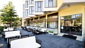 An outdoor seating area with multiple tables and chairs, a building with large windows, and a paved walkway.