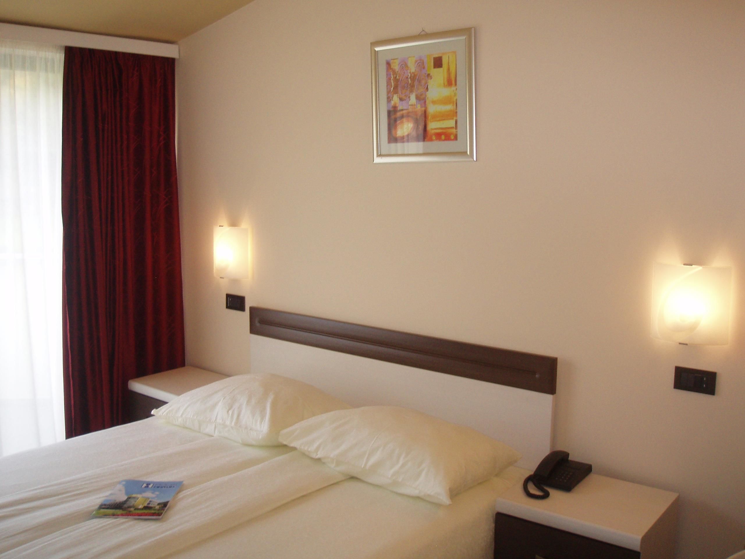 A hotel room with a bed, two bedside tables, a nightstand, a telephone, and a painting on the wall.