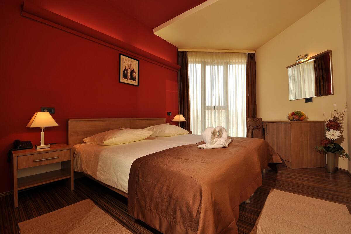 A hotel room with a large bed, two bedside tables with lamps, a wooden dresser, a mirror, and a window with curtains.