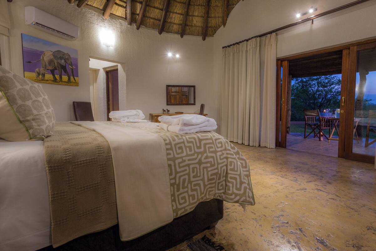 A well-lit bedroom with a bed, a nightstand, and a painting of elephants. There is a sliding glass door leading to an outdoor area with a table and chairs.