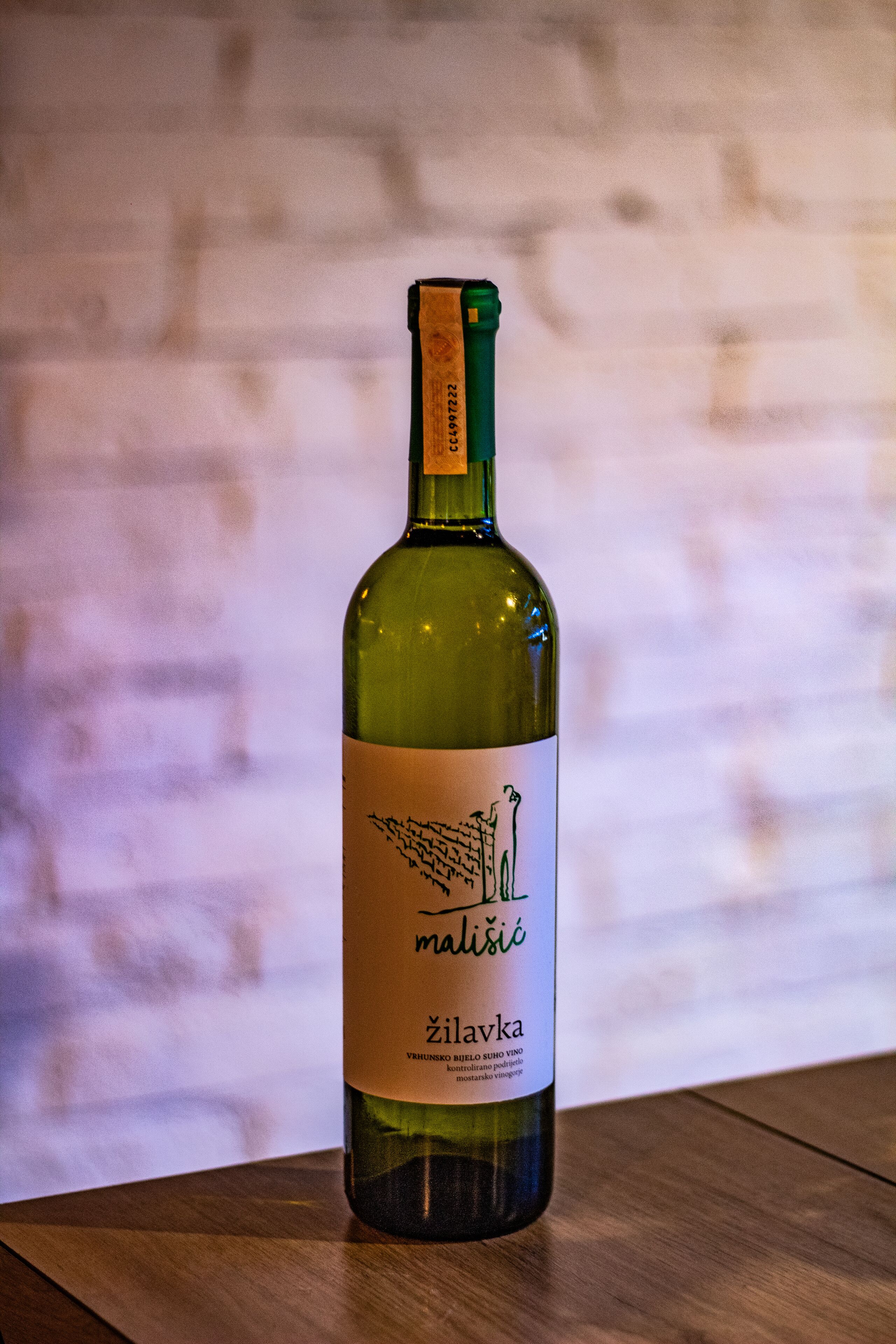 Una botella de vino Zilavka malvísci sobre una superficie de madera frente a un muro de ladrillo.