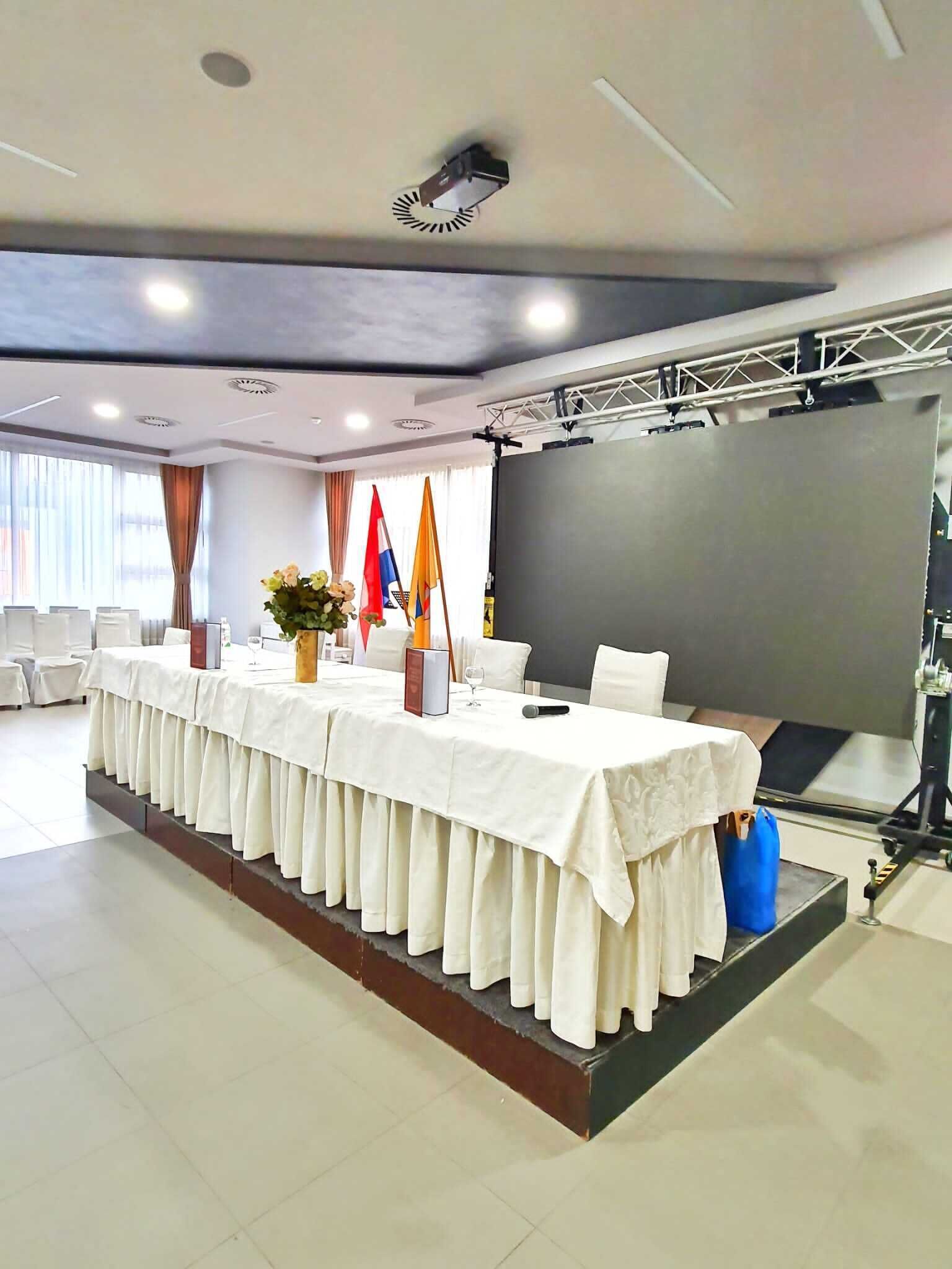 Une salle de conférence avec une longue table, des drapeaux, un projecteur et un écran.