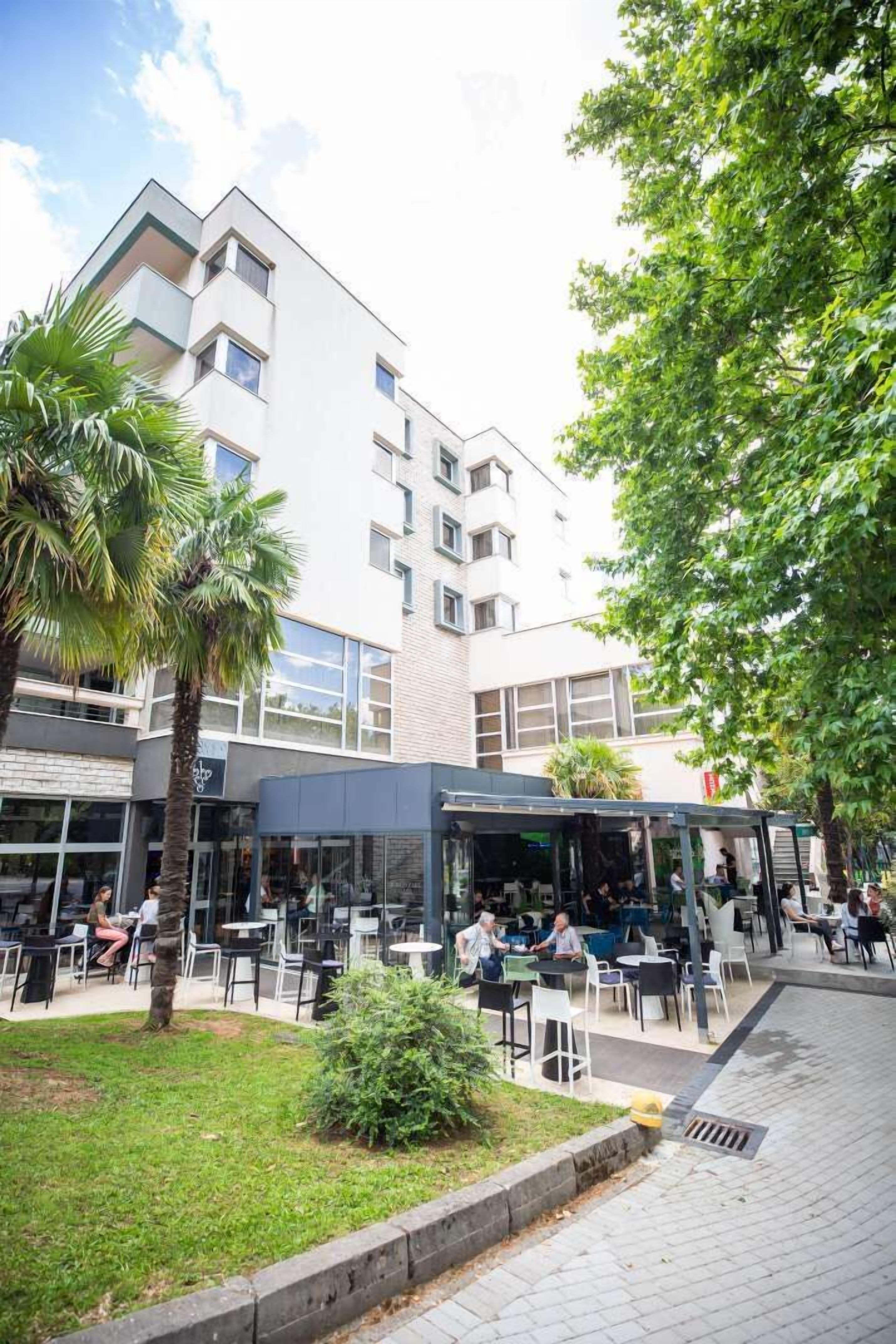 Un espacio al aire libre con mesas y sillas, rodeado de follaje y un edificio de varios pisos y ventanas.