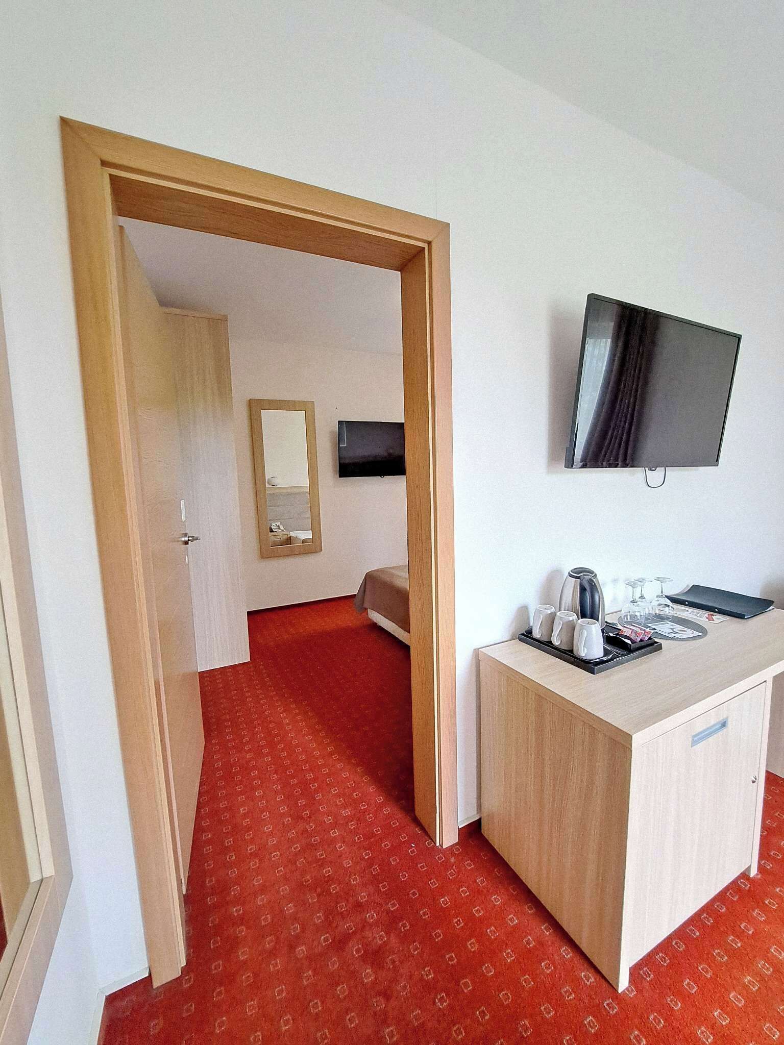 Camera d'albergo con tappeto rosso, una scrivania in legno con bollitore, tazze e una rivista, un letto, una TV a parete e uno specchio sulla porta.
