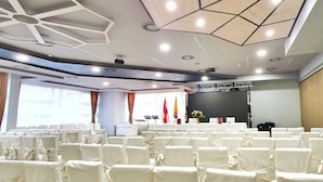 Un salón de conferencias con un escenario, filas de sillas con fundas blancas y un diseño geométrico en el techo.