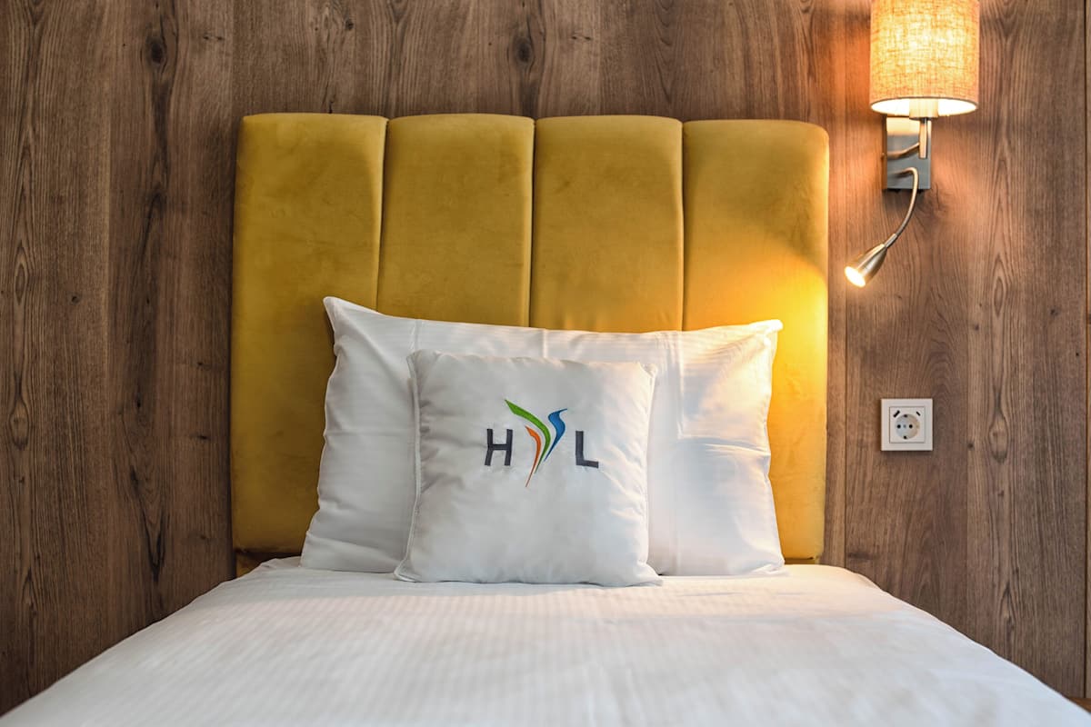 Una cama con cabecera amarilla, almohada blanca con un logo, y una lámpara fijada a la pared.