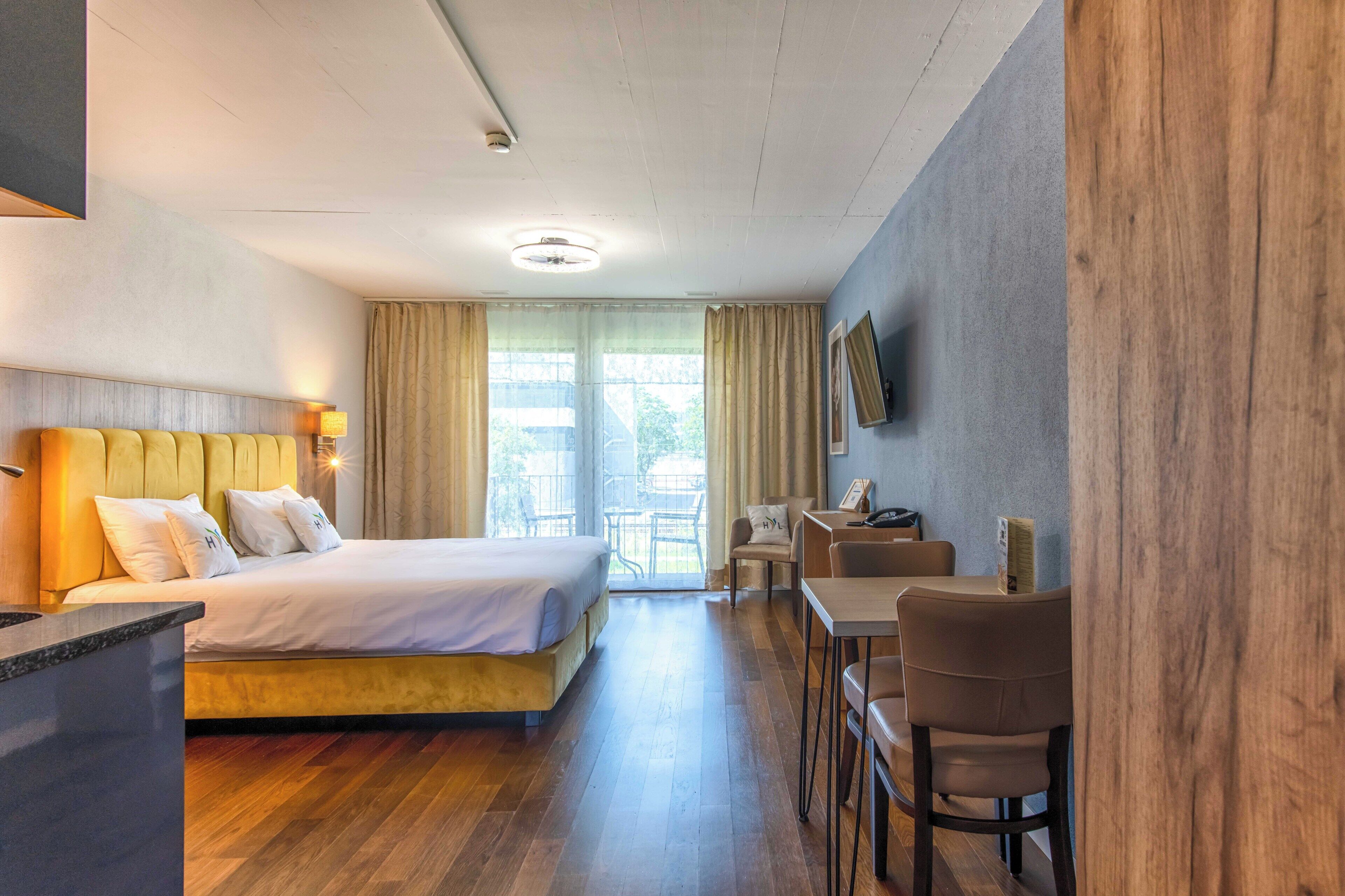 Hotellihuone, jossa on sänky, työpöytä ja tuoli. Huoneessa on ikkuna, jossa on verhot, ja ikkunasta on näkymä ulos.