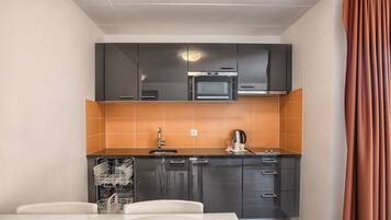 Cocina compacta con microondas, horno y salpicadero de azulejos naranjas. Incluye lavavajillas y una mesa blanca con sillas.