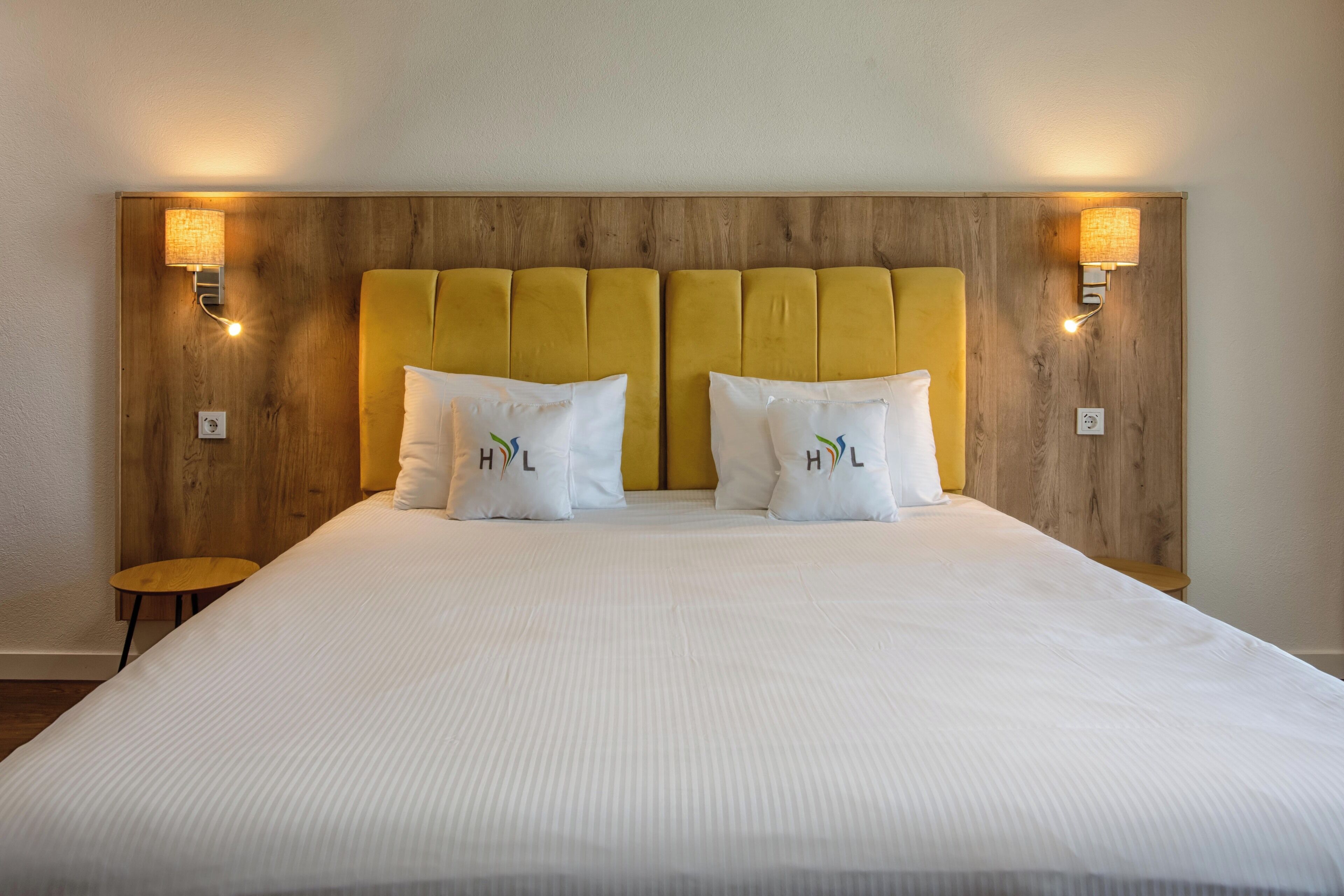 Una habitación de hotel con cabecera de madera, dos mesitas de noche y una cama con ropa de cama y almohadas blancas.