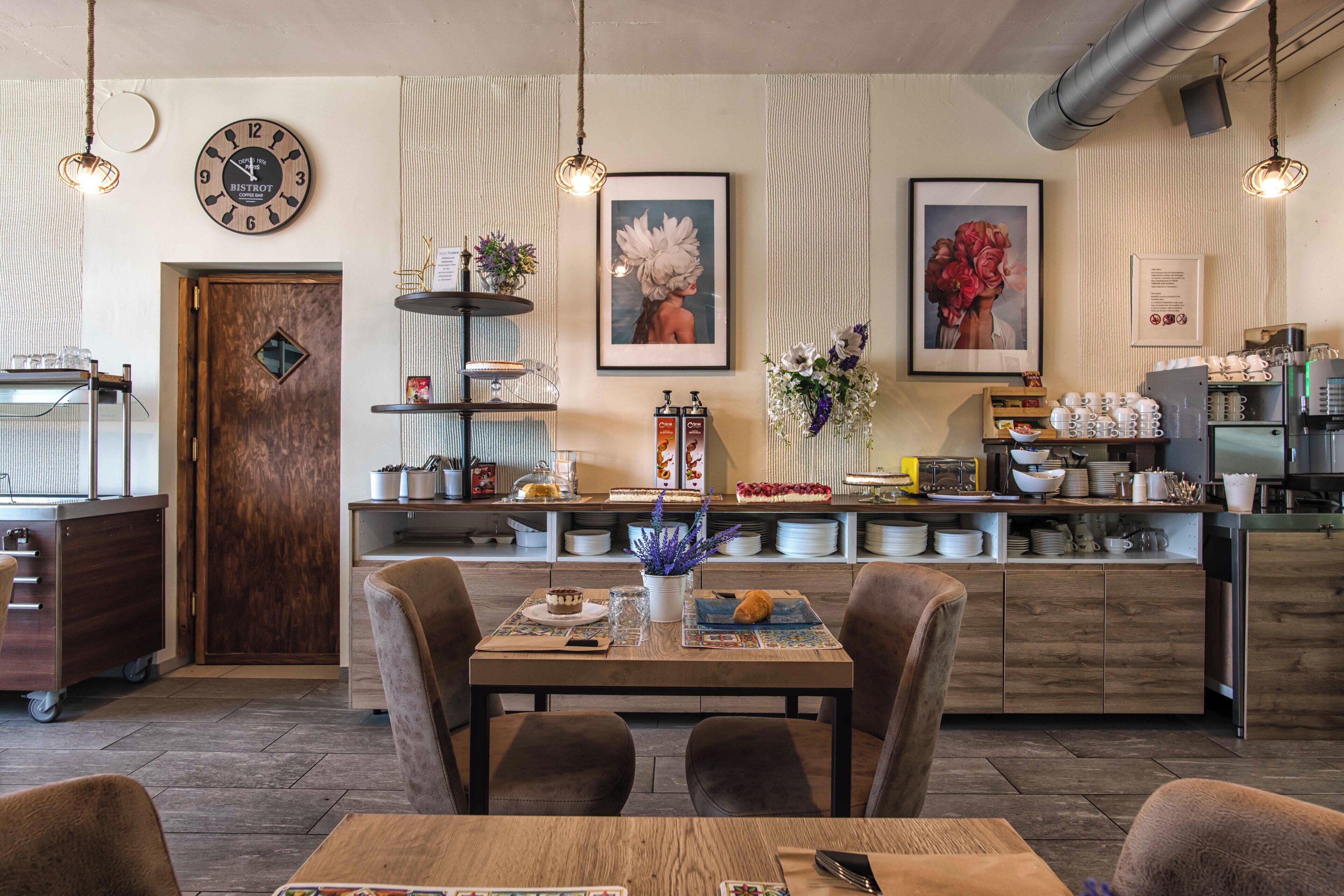 L’intérieur d’un restaurant, avec une table en bois, des chaises, une horloge, des œuvres d’art encadrées et un comptoir de cuisine où sont disposés divers objets.