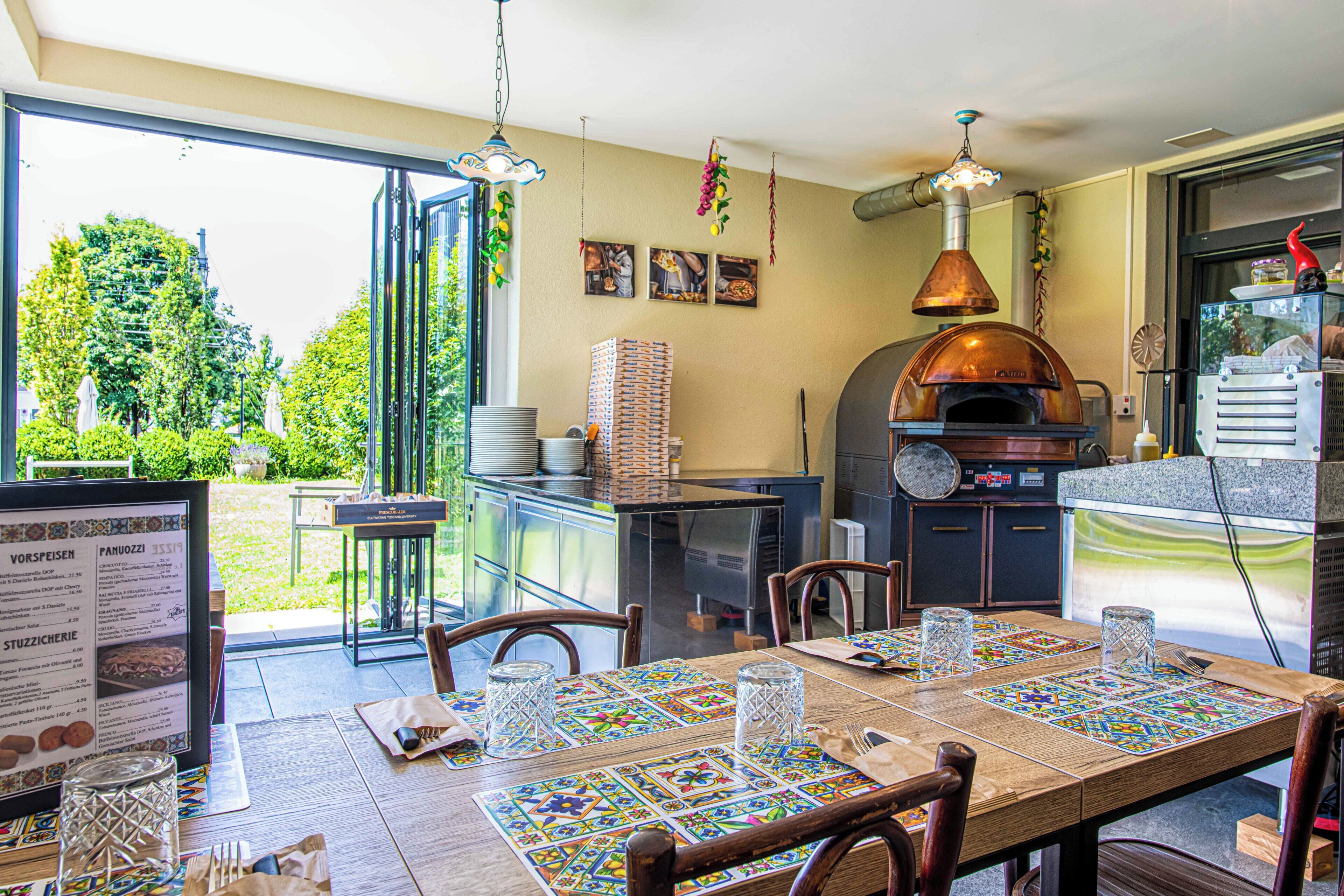 Eine offene Wohnküche mit Essbereich, ein großes Fenster, ein Pizzaofen und ein Esstisch, der mit Platzsets und Gläsern gedeckt ist.