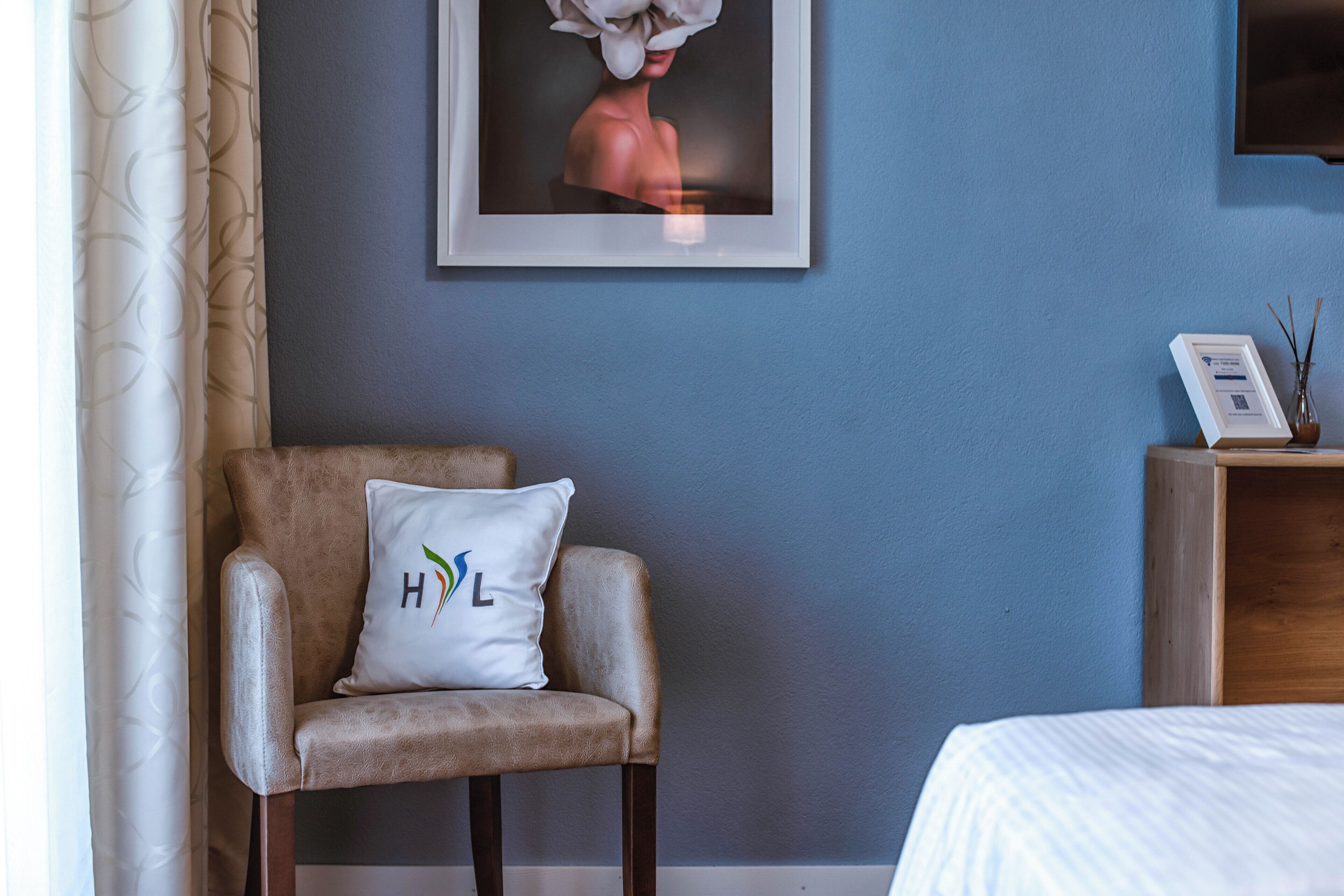 Ein Zimmer mit einem braunen Sessel, einem weißen Kissen mit einem Logo, einem gerahmten Kunstwerk an der Wand und einem Holznachttisch mit einer Vase.